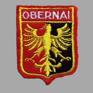 May include: Embroidered patch with a red border and a yellow inner border. The patch features the word "OBERNAI" in white letters at the top. Below, a yellow eagle is centered against a red and black background.