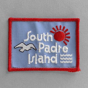 May include: Embroidered patch featuring the words "South Padre Island" in white script on a light blue background. A red sun and a white seagull are also embroidered on the patch, which has a red border.