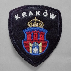 May include: A shield-shaped embroidered patch with a black background and the white text KRAKÓW at the top. The center features a gold crown above a blue and red crest depicting a brick wall with three towers and a white eagle emblem at the base.