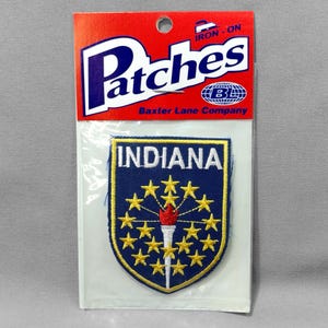 May include: An iron-on patch featuring the state of Indiana. The patch is blue with a gold border and features a torch and 13 stars. The text "INDIANA" is printed in white on the patch. The patch is packaged in a clear plastic bag with a red, white, and blue header that reads "Patches Baxter Lane Company".