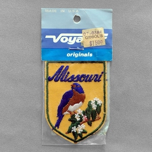 May include: A Missouri state patch featuring a bluebird perched on a flowering branch, embroidered on a yellow shield. The word "Missouri" is embroidered in blue script. The patch is packaged in a clear plastic bag with a blue and white header that reads "Voya originals."