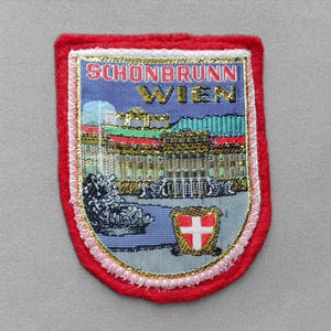 May include: A rectangular embroidered patch with a red felt border featuring a depiction of Schönbrunn Palace in Vienna. The text SCHÖNBRUNN WIEN is stitched in red and gold thread above the palace, with a small shield featuring a white cross.