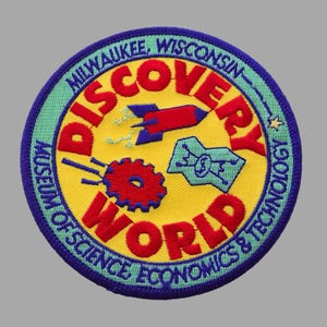 May include: Embroidered patch with a yellow center and a blue border. The patch features the words "DISCOVERY WORLD" in red, with illustrations of a rocket, gear, and circuit board. Text around the border reads "MILWAUKEE, WISCONSIN MUSEUM OF SCIENCE ECONOMICS & TECHNOLOGY."