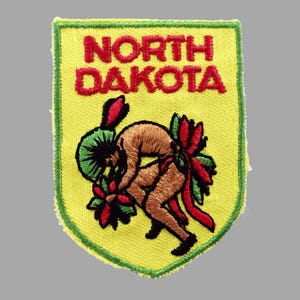 May include: Embroidered patch featuring the words "NORTH DAKOTA" in red letters. The patch has a yellow background with a green border. The design includes a brown figure with green and red floral accents.