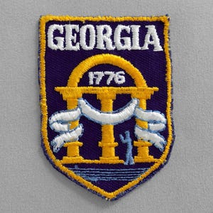 May include: A purple and gold embroidered patch with the word "GEORGIA" in white lettering and the year "1776" in white lettering. The patch features a white embroidered archway with a white ribbon and a blue figure standing in the center.