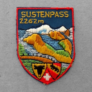 May include: A blue embroidered patch with the text "SUSTENPASS 2262m" and a depiction of a mountain range and a lake. The patch features a red border and a Swiss flag.