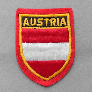 May include: A red, white, and yellow embroidered patch with the word "AUSTRIA" in gold letters. The patch is shield-shaped and features the Austrian flag design.