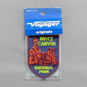 May include: A blue patch with the text "BRYCE CANYON NATIONAL PARK" in yellow and orange. The patch features an embroidered image of the Bryce Canyon rock formations in orange. The patch is in a clear plastic sleeve and has a blue header that says "Voyager originals" and "MADE IN U.S.A."