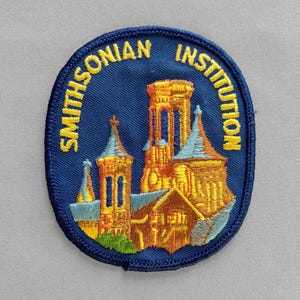 May include: Embroidered patch with the words "Smithsonian Institution" in yellow around a blue oval. The patch features a detailed illustration of a building with multiple towers in shades of gold, orange, and blue.