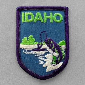 May include: Embroidered patch featuring the word "IDAHO" in green letters above a blue background. The patch depicts a fish jumping out of the water, with green and purple details. The patch has a purple border.