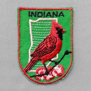 May include: Embroidered patch featuring a red cardinal perched on a branch with red and white flowers. The patch is green with a red border and the word "INDIANA" at the top. A white outline of the state is behind the bird.