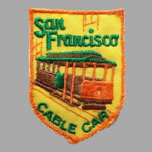 May include: A yellow embroidered patch with an orange border. The patch features a cable car with a brown body and orange windows. The text "San Francisco Cable Car" is embroidered in green on the patch.