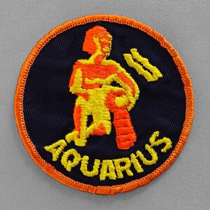 May include: A round, navy blue fabric patch with an orange border. The patch features a yellow and orange embroidered image of the astrological sign Aquarius. The text "Aquarius" is embroidered in yellow below the image.