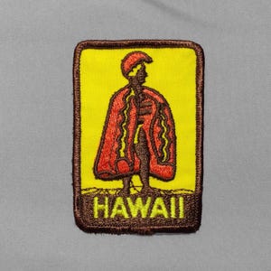 May include: A rectangular embroidered patch with a yellow background and a dark brown border. It features a figure wearing a red cloak and a helmet, standing above the word HAWAII stitched in yellow capital letters against a brown base.