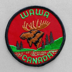 May include: Embroidered patch with a red background, featuring a moose and the words "WAWA CANADA" in green. The moose is brown and black, with green and black trees and a river at the bottom. The patch has a black border.