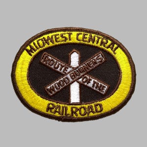 May include: An embroidered patch with a yellow and brown oval border. The patch features the text "MIDWEST CENTRAL RAILROAD" and "ROUTE WOOD BURNERS OF THE" in a circular design. A white cross is in the center.