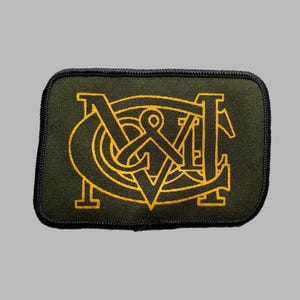 May include: A rectangular patch with a dark green background and a gold embroidered design. The design features stylized letters and symbols, with a black border. The patch is likely for clothing or accessories.