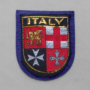 May include: A patch with the word "ITALY" in gold lettering. The patch features a shield design with a gold lion, a red and silver cross, and a black and silver cross. The patch is bordered with blue and gold trim.