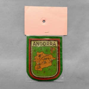May include: A rectangular felt patch featuring a map of Andorra in orange on a green background with a red border. The word ANDORRA is printed in black at the top. The patch is enclosed in a clear plastic sleeve attached to a light pink paper header.