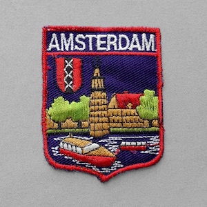 May include: A rectangular embroidered patch with a red border featuring the word AMSTERDAM in white text. The design depicts a blue background with a red shield containing three white crosses, a tall tan tower, green trees, and a red boat on water.