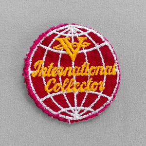 May include: Embroidered patch featuring a red globe design with white grid lines. The patch has the words "International Collector" in yellow script. A yellow "V" is at the top. The patch has a pink border and is on a gray background.