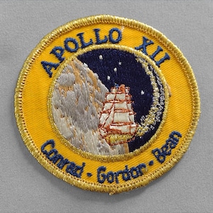 May include: A yellow embroidered patch with gold trim featuring the text "Apollo XII Conrad - Gordon - Bean" and a depiction of a ship sailing past a crescent moon.