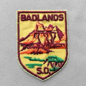 May include: A shield-shaped embroidered patch with a yellow background and a maroon border. It features the text BADLANDS at the top and S.D. at the bottom in maroon. The center depicts a landscape with maroon and orange rock formations and green.