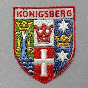 May include: Embroidered patch featuring the word "KÖNIGSBERG" at the top. The patch has a shield design with a red border, and includes crowns, stars, and a cross in various colors.