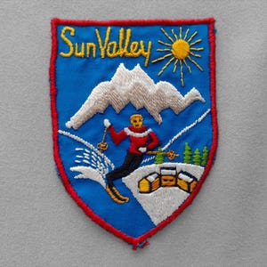 May include: A shield-shaped embroidered patch with a blue background and a red border. It features the yellow text Sun Valley, a yellow sun, white mountains, a skier in a red jacket and black pants, and small yellow buildings on snow.