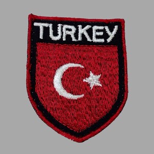 May include: Embroidered patch featuring the word "TURKEY" in white letters above a red shield. The shield displays a white crescent moon and star, symbols of Turkey. The patch has a black border and a red stitched edge.