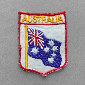 May include: A shield-shaped embroidered patch featuring a white background with a red border. The word AUSTRALIA is stitched in yellow at the top. Below, a yellow flagpole holds a blue Australian flag with white stars and a Union Jack.