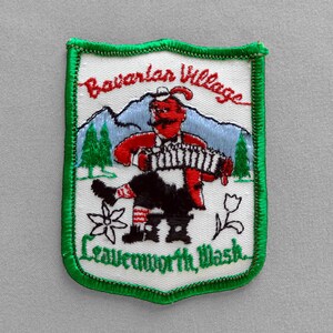 May include: Embroidered patch with a green border and a white background. The patch features a red figure playing an accordion, mountains, and the text "Bavarian Village Leavenworth Wash."