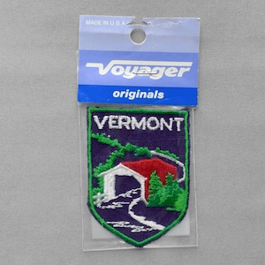 May include: A shield-shaped embroidered patch featuring a covered bridge with a red roof, green trees, and a winding path on a dark blue background. The word VERMONT is stitched in white at the top. The patch is in a clear plastic package labeled VOYAGER originals.