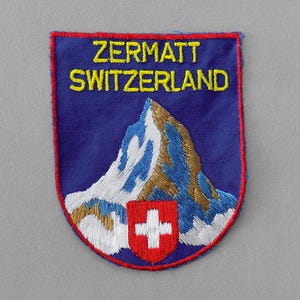 May include: Embroidered patch featuring the text "ZERMATT SWITZERLAND" in yellow, above a mountain illustration. The patch has a blue background, a red border, and a Swiss flag emblem.