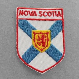 May include: A white embroidered patch with the words "NOVA SCOTIA" in red lettering. The patch features a blue and white cross with a yellow shield containing a red lion in the center.
