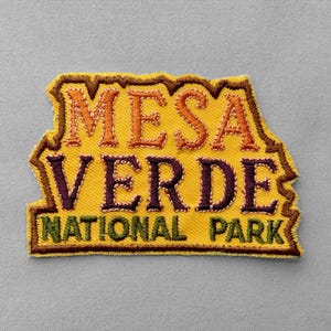 May include: An embroidered patch with a yellow background and a dark brown border. The text MESA VERDE is stitched in orange and dark purple, while the words NATIONAL PARK are stitched in dark green. The patch has an irregular, jagged shape.