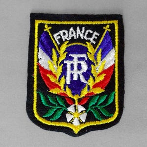 May include: Embroidered patch featuring the word "FRANCE" in white, arched above a central emblem. The emblem includes a blue and white design, flanked by flags in red, white, and blue. The patch has a black background with yellow and green accents.