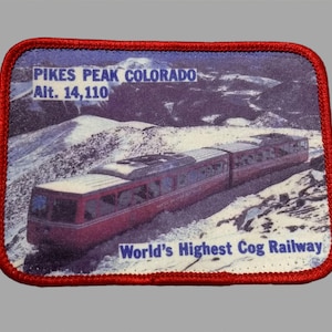 May include: Embroidered patch featuring a red cog railway train ascending a snowy mountain. The patch reads "Pikes Peak Colorado Alt. 14,110" and "World's Highest Cog Railway" in white text. The patch has a red border.