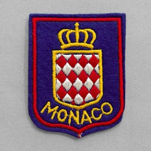 May include: A rectangular patch with a shield design. The shield is blue with a red border and features a gold crown above a red and white checkered pattern. The word "MONACO" is embroidered in gold.