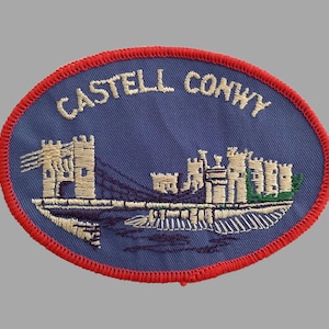 May include: Embroidered patch with a blue oval background and a red border. The patch features the text "CASTELL CONWY" in white, above an image of a castle and bridge in white and green.