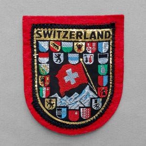 May include: A red felt patch featuring the word SWITZERLAND in gold thread at the top. The center displays a red flag with a white cross above stylized mountains, surrounded by a border of small, colorful embroidered heraldic shields.