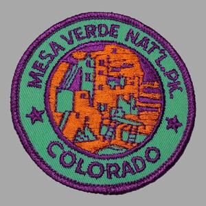 May include: A purple and green embroidered patch with the text "Mesa Verde Nat'l Pk. Colorado" and an orange silhouette of a cliff dwelling.