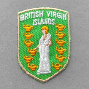 May include: An embroidered shield-shaped patch featuring a green background with the text BRITISH VIRGIN ISLANDS in white. The design includes a central white figure in a robe surrounded by twelve small yellow oil lamps arranged in two columns.