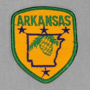 May include: A green and yellow embroidered patch with the word "ARKANSAS" in green letters. The patch features a blue outline of the state of Arkansas with a pine tree inside. There are five blue stars around the state outline.