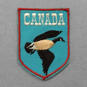 May include: Embroidered patch with a teal background and a red border. The patch features the word "CANADA" in white block letters and a flying goose with black wings and a tan body.