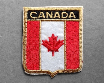May include: A rectangular embroidered patch featuring the word CANADA in gold thread on a black background above a shield design. The shield displays a red maple leaf centered on a white vertical stripe flanked by two red stripes, all bordered in gold.
