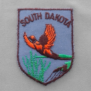 May include: Embroidered patch with a blue background and brown border. The patch features a flying pheasant with orange and brown feathers, green grass, and the text "SOUTH DAKOTA" in brown letters.