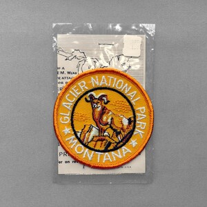 May include: A circular embroidered patch featuring a bighorn sheep standing on rocks against a yellow background. The border is orange with the white text Glacier National Park Montana. The patch is enclosed in a clear plastic sleeve with a paper insert.
