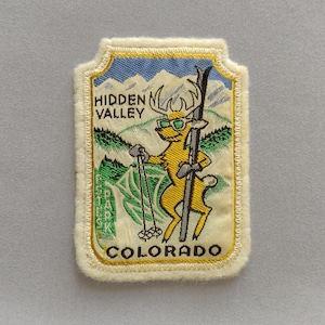 May include: A rectangular embroidered patch with a cream-colored border featuring a yellow deer wearing green sunglasses and holding a ski pole and a single ski. The background depicts blue mountains and green trees with the text HIDDEN VALLEY COLORADO.