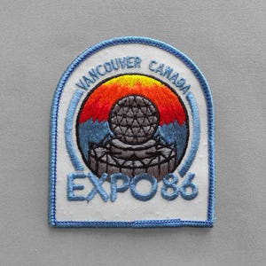 May include: Embroidered patch featuring the Vancouver Canada EXPO 86 logo. The design includes a colorful sunset over a geodesic dome, with text in blue. The patch has a white background with a blue border.